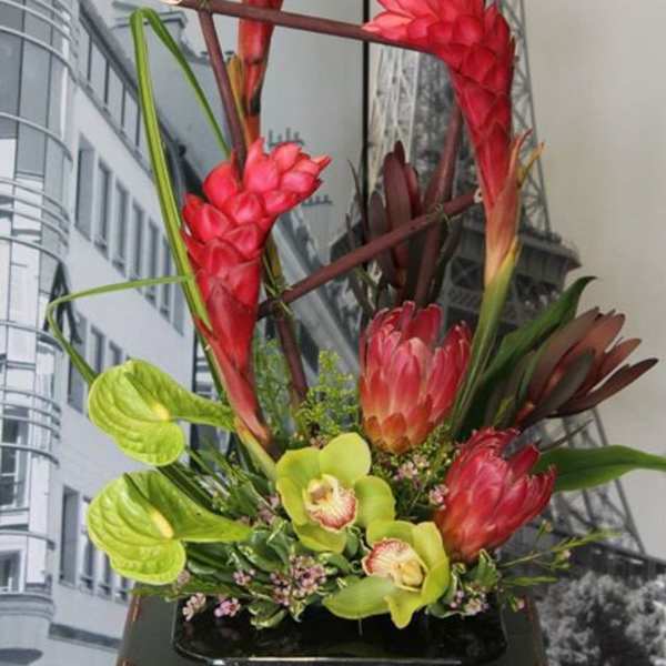 Tall modern arrangement of red tropical flowers, pink protea, and green orchids in a low black container