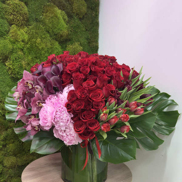 Large arrangement of red roses, pink peonies, orchids and tulips in a glass vase with broad tropical leaves.