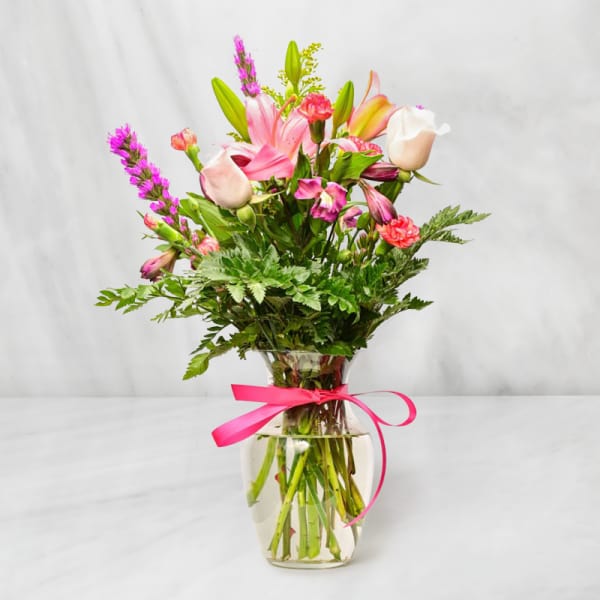 Mixed bouquet of pink lilies, roses, and carnations in a clear glass vase with a pink ribbon