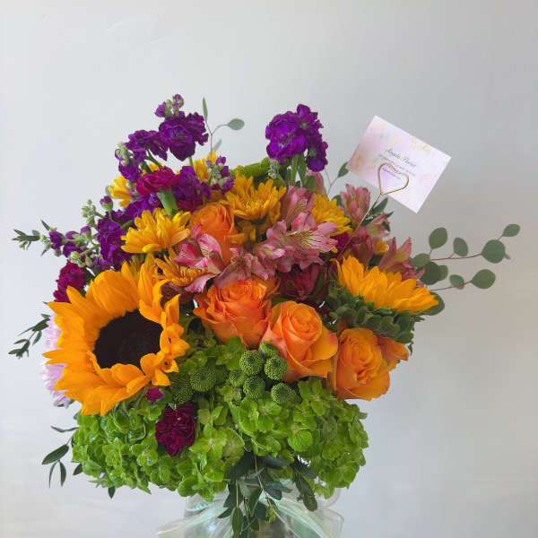 Bouquet of sunflowers, orange roses, and purple blooms in a glass vase