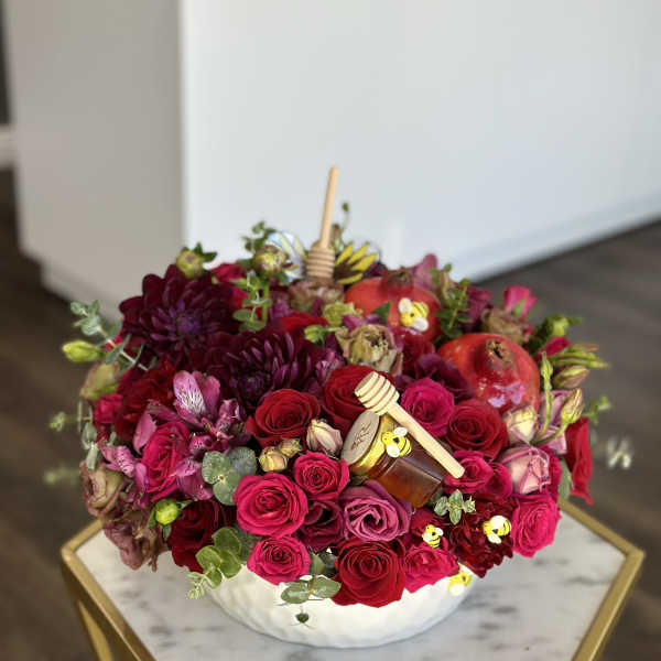 Round arrangement of red and pink roses with honey jars and bee picks in a white bowl