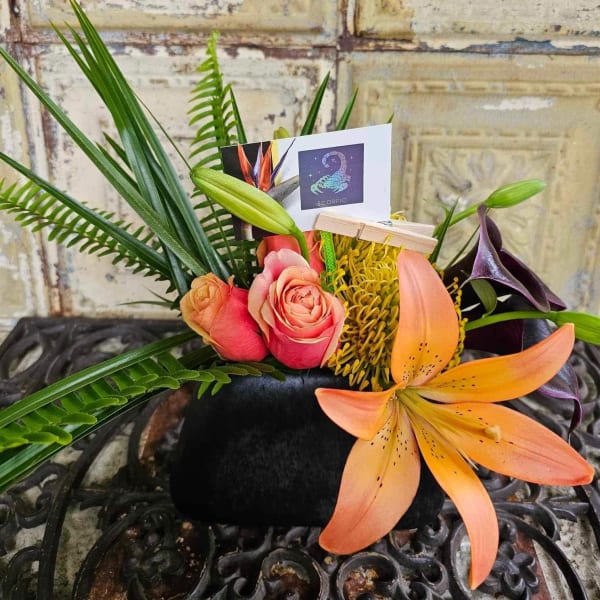 Tropical floral arrangement with roses, lilies, and a Scorpio card in a black container