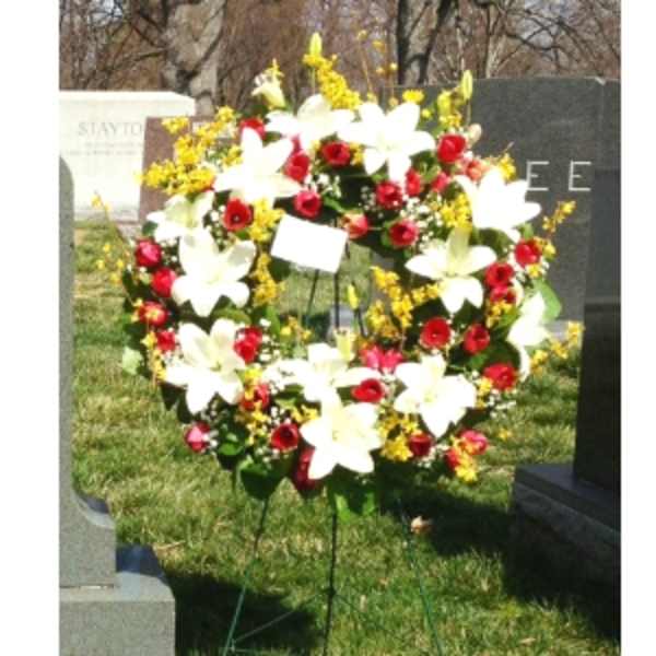 Standing wreath of white lilies, red roses, and yellow flowers on an easel in a cemetery.