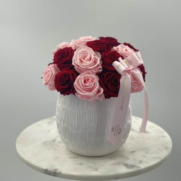 Pink and red roses arranged in a white vase with a pink ribbon