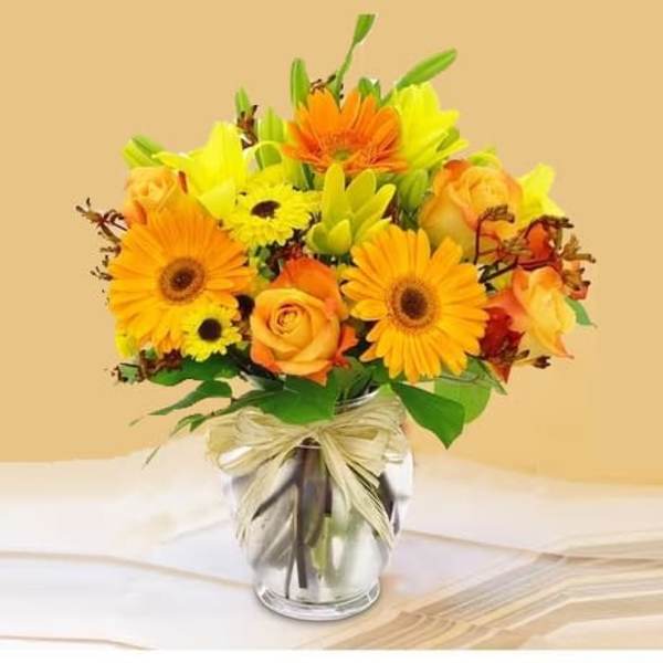 Bright orange gerberas, yellow lilies, and roses in a clear glass vase with a raffia bow