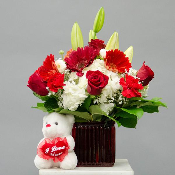 Red and white flower arrangement in a glass vase with a teddy bear