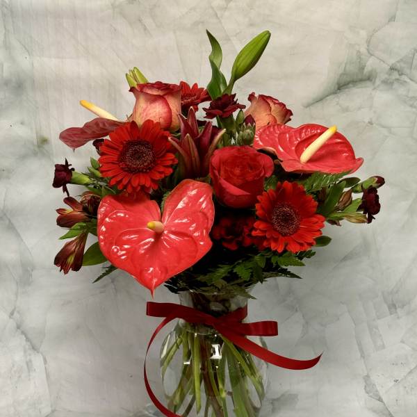 Red floral bouquet with anthuriums, roses, and gerbera daisies in a glass vase