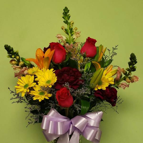 Mixed bouquet of roses, lilies, and yellow daisies in a glass vase with a lavender bow