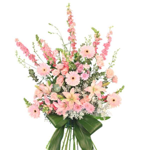 Tall standing spray of pink lilies, gerbera daisies, carnations and snapdragons with large green leaves on an easel