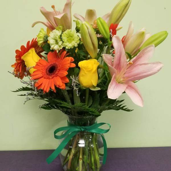 Mixed bouquet of lilies, roses, and gerbera daisies in a glass vase with ribbon