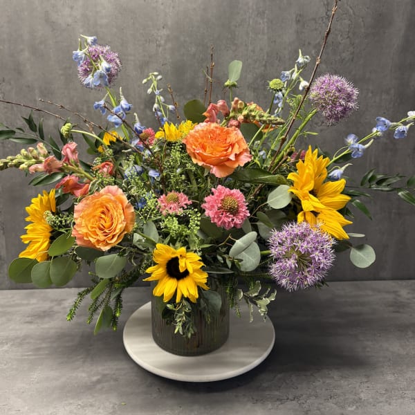 Mixed bouquet with orange roses, sunflowers, and purple allium in a glass vase