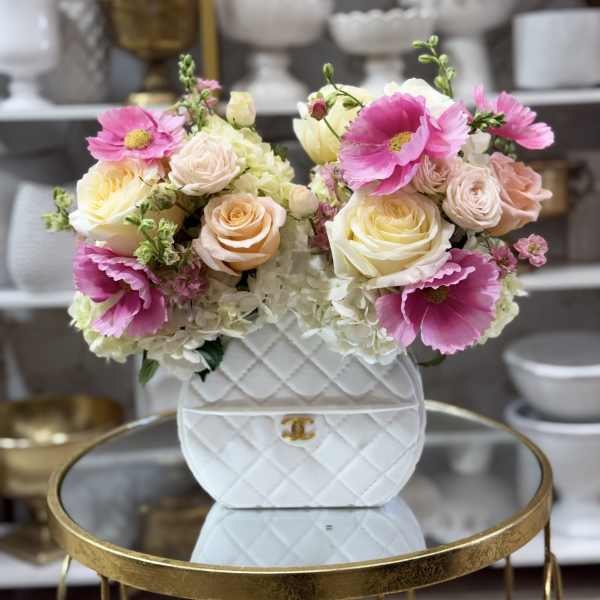 Handbag-shaped white ceramic vase filled with pink anemones, pastel roses, and white hydrangeas on a gold table