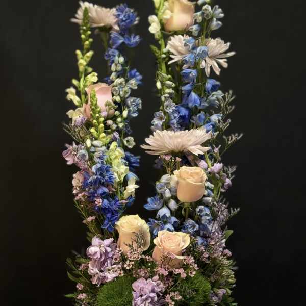 Tall pastel arrangement of peach roses and blue delphinium in a white bowl vase on draped fabric