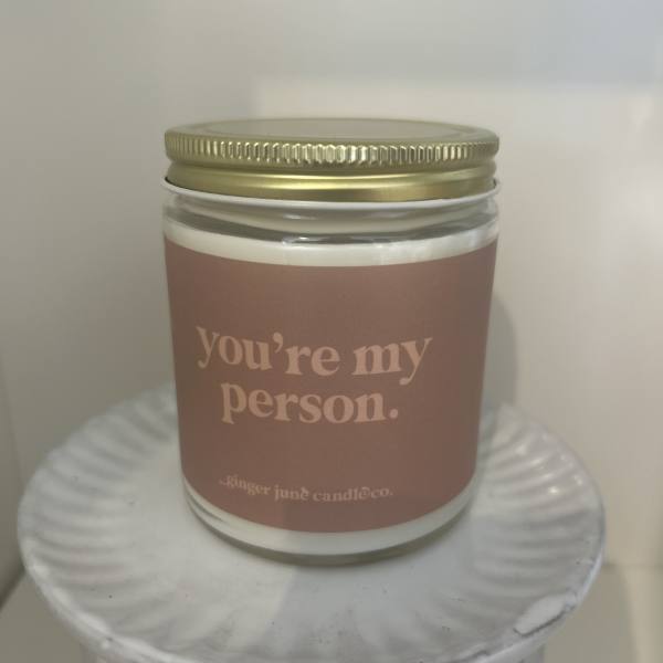 Glass jar candle with gold lid and label reading "you’re my person" on a white pedestal stand.
