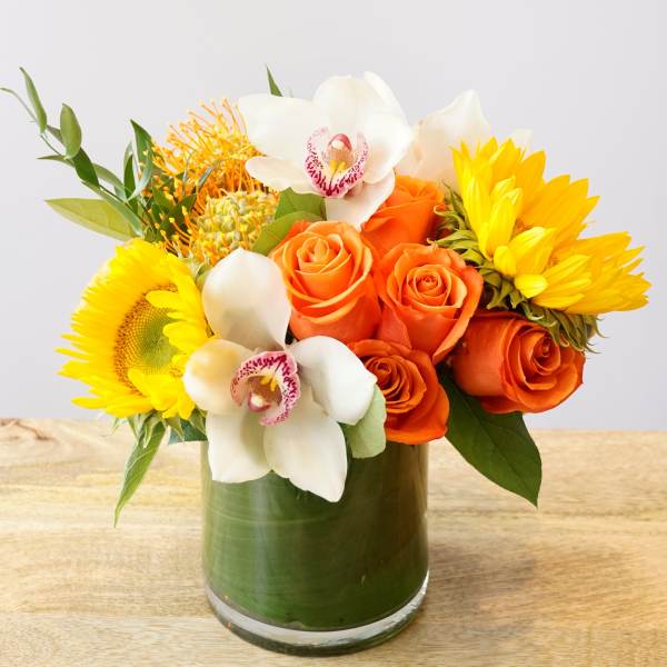 Orange roses and white orchids with yellow sunflowers in a glass vase