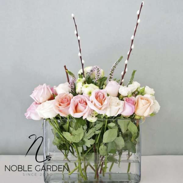 Pink and white roses in a clear rectangular vase with tall pussy willow stems