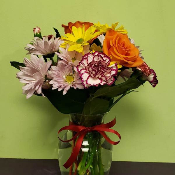 Mixed bouquet of daisies, roses, and carnations in a glass vase with a red ribbon