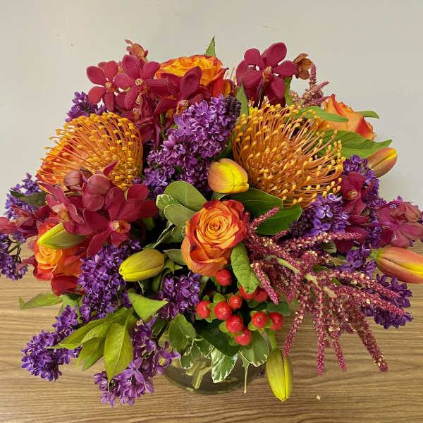 Bright mixed bouquet with orange roses, purple blooms, and yellow tulips in a glass vase