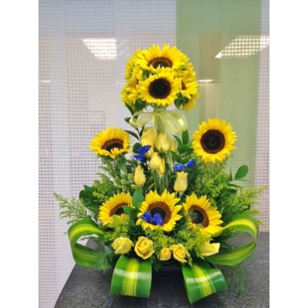 Tall sunflower and yellow rose arrangement with blue butterfly accents and green ribbon in a low container