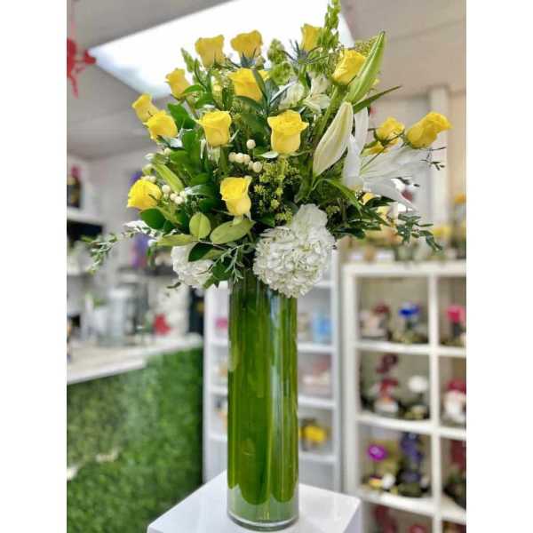 Tall arrangement of yellow roses, white lilies, and hydrangeas in a green-lined glass vase