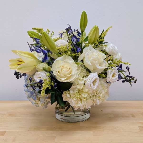 White roses, lilies, and hydrangeas with blue accents arranged in a low clear glass vase