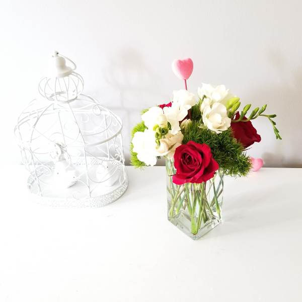 Red and white flowers arranged in a clear square vase beside a white birdcage