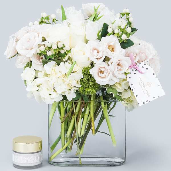White roses and hydrangeas arranged in a clear glass vase with a small candle jar beside it.
