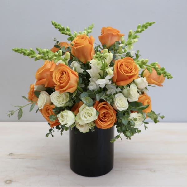 Orange and ivory roses with white snapdragons arranged in a black cylinder vase