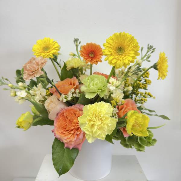 Bright arrangement of yellow gerbera daisies, peach roses, and mixed blooms in a white cylinder vase