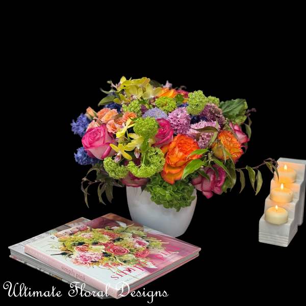 Colorful mixed flower arrangement in a white vase beside candles and books