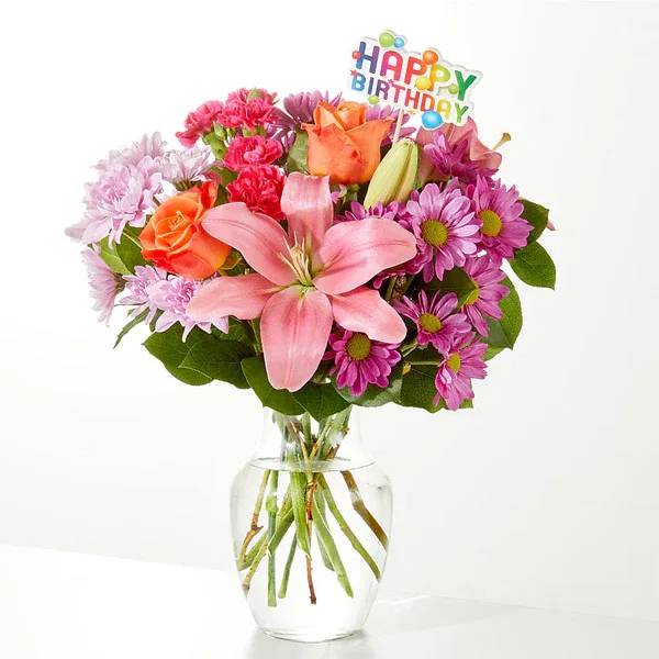 Pink and orange mixed bouquet in a clear glass vase with a birthday topper