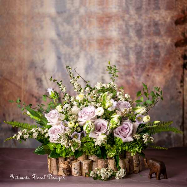 Low arrangement of lavender roses and white blooms in a rustic birch container with a small wooden elephant.