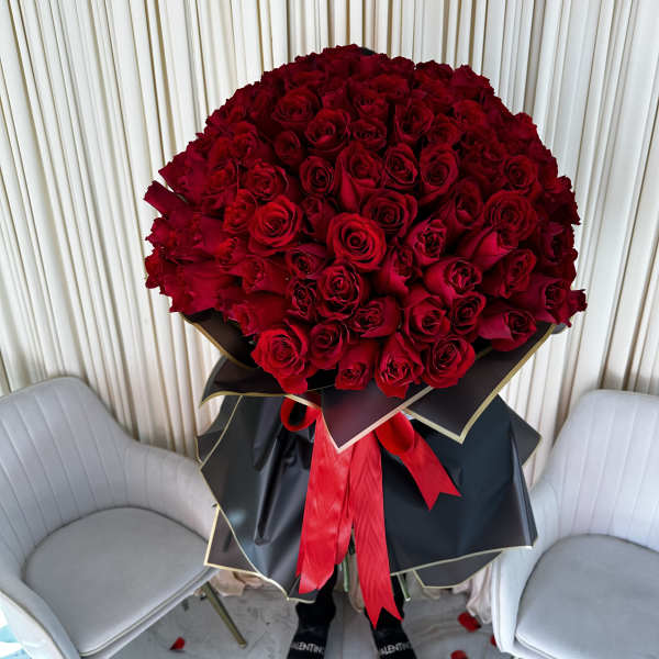 Oversized bouquet of red roses wrapped in black paper with gold trim and a red ribbon, held between two white chairs