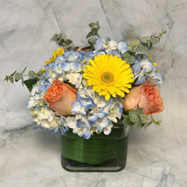 Bouquet of blue and white hydrangeas with yellow gerbera daisies and peach roses in a glass vase