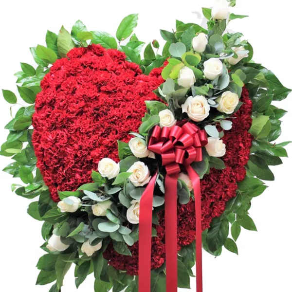 Standing red carnation heart spray with white roses and red ribbon on a greenery backing