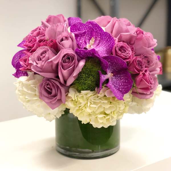 Low round arrangement of mauve roses, purple orchids, and white hydrangeas in a clear glass cylinder vase