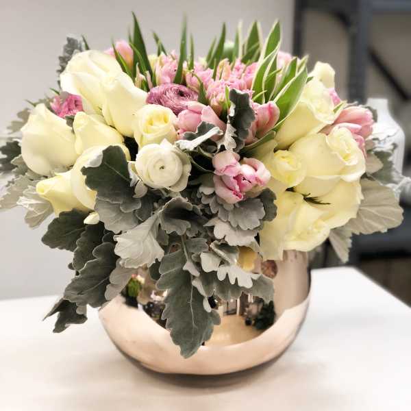 Low arrangement of ivory roses, pink tulips, and ranunculus in a round metallic bowl.