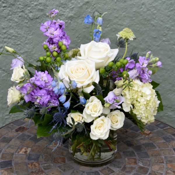 White roses, hydrangea, and purple stock arranged with blue accents in a clear glass vase on a mosaic table