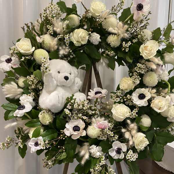 White floral wreath on an easel with a plush teddy bear in the center