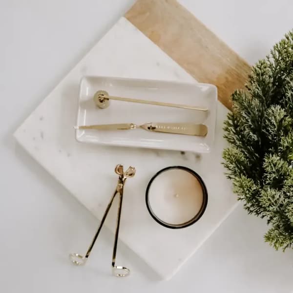 Gold candle care tools with small white candle and faux evergreen on marble boards