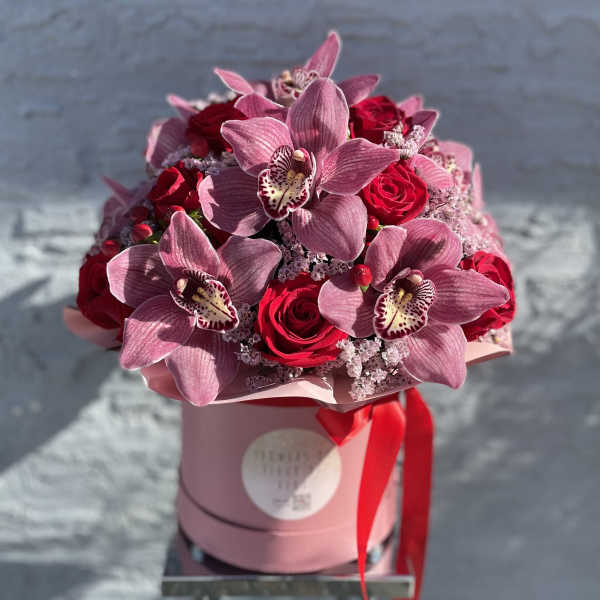 Pink orchids and red roses arranged in a round hatbox with a red ribbon
