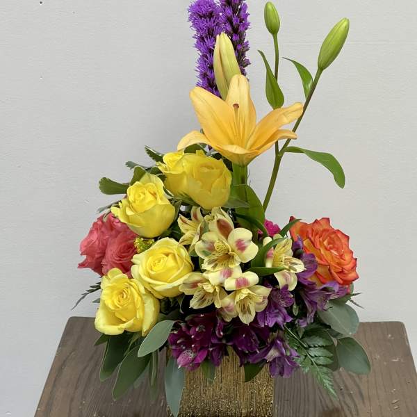Mixed bouquet with yellow roses, lilies, and purple flowers in a gold vase