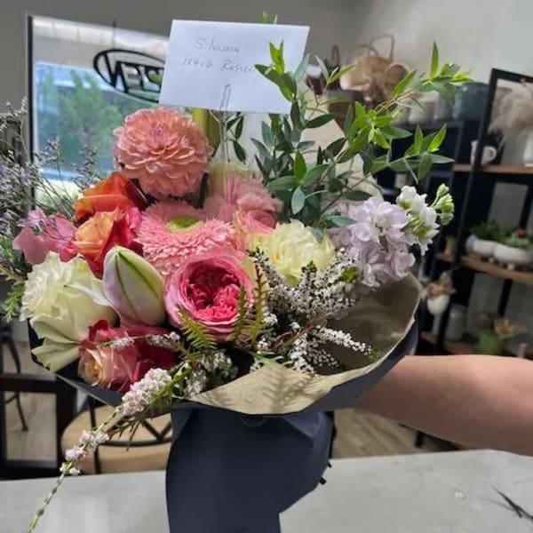 Hand-tied bouquet of pink, peach, and white flowers wrapped in dark paper