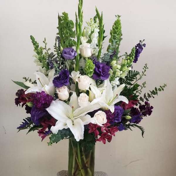 Tall floral arrangement with white lilies, roses, and purple blooms in a glass vase