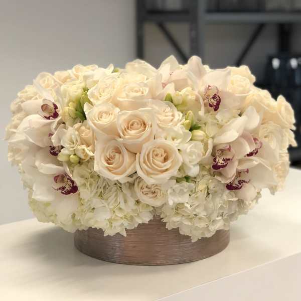 Low round arrangement of cream roses, hydrangeas, and orchids in a short metallic container