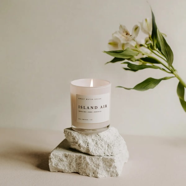 Lit candle in a glass jar beside a white flower stem on stacked stones