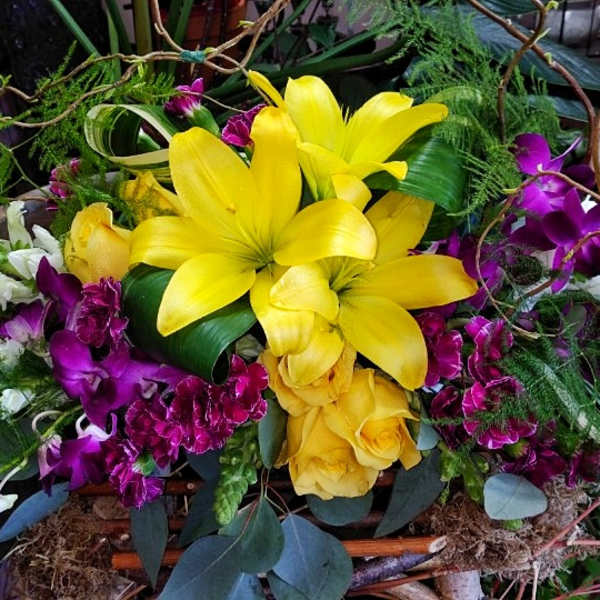 Bright yellow lilies and purple orchids arranged with roses in a decorative basket.