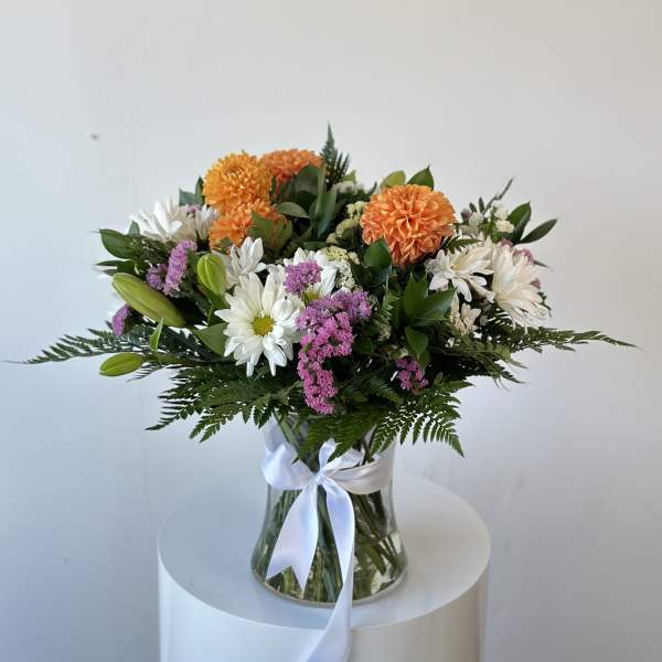 Mixed bouquet of orange, white, and purple flowers in a glass vase with a white ribbon