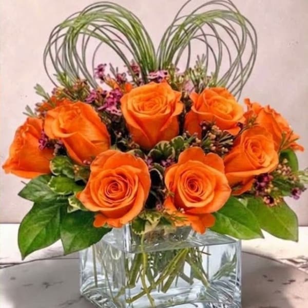 Orange roses in a clear square vase with heart-shaped wire accents