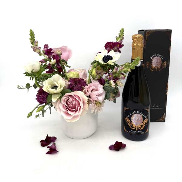 Pink and white floral arrangement in a white vase with a bottle of sparkling wine and gift box.